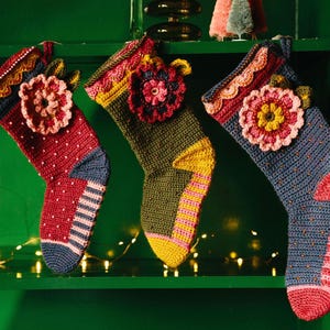 May include: Three crocheted Christmas stockings with floral designs in shades of pink, red, blue, green, and yellow hanging on a green shelf.