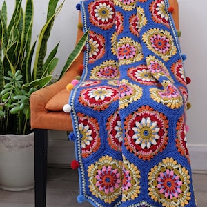May include: A crocheted throw blanket with a blue background and colourful floral patterns. The blanket is decorated with pom-poms along the edges and is draped over a chair.