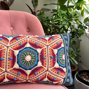 以下が含まれることがあります： 赤、オレンジ、青、クリーム色を基調とした、鮮やかな幾何学模様の長方形のかぎ針編みクッション。ピンクの椅子の上に置かれ、背景には緑豊かな植物が見えます。