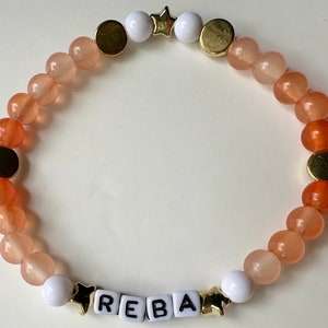 May include: A peach and orange ombre beaded bracelet with gold spacers and white star charms. The bracelet has the name "REBA" spelled out in white letter beads.