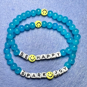May include: Two beaded bracelets with turquoise beads. One bracelet has the words "BE HAPPY" spelled out in white letter beads, and the other says "SPARK JOY". Both bracelets have yellow smiley face beads.
