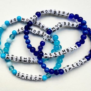 May include: Three beaded bracelets with blue and turquoise beads. The bracelets spell out the words "FREEDOM", "ASSEMBLY", and "SPEECH".