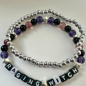 May include: Two beaded bracelets. The top bracelet features silver beads and a mix of purple, black, and pink beads. The bottom bracelet spells "RAGING WITCH" with black and white letter beads and silver star accents. The bracelets are on a white background.