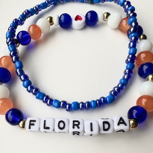 May include: Two beaded bracelets with blue, white, and orange beads. The bottom bracelet has white letter beads spelling out "FLORIDA".