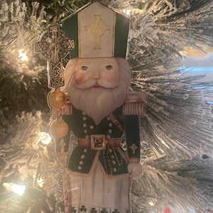 May include: A green and white Christmas ornament depicting a figure in a green uniform holding a staff with a golden orb. The figure wears a white hat with a green trim and a gold cross. The ornament has a Celtic cross design.