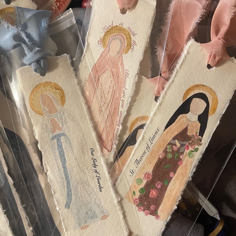 Może przedstawiać: Cztery ręcznie robione zakładki z postaciami religijnymi. Zakładki są wykonane z papieru o fakturowanej powierzchni i przedstawiają ręcznie malowane obrazy Matki Boskiej z Lourdes, Świętej Teresy od Dzieciątka Jezus i dwie inne postacie. Zakładki zawierają tekst: "Pan jest z tobą. Błogosławiona jesteś między niewiastami. Matka Boska z Lourdes. Święta Teresa od Dzieciątka Jezus."