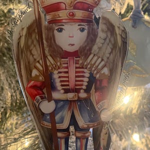May include: A Christmas ornament featuring a nutcracker angel with wings, holding a staff. The angel wears a red and blue uniform with a crown. The ornament is part of the Nutcracker Collection.