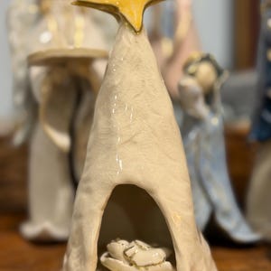 May include: A cream-colored ceramic figurine shaped like a conical tent with a textured, crackled finish. A bright yellow star sits at the peak. Inside the open arched doorway, a small, detailed figure rests on a base with gold accents.