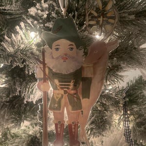 May include: A vintage-style Christmas bauble depicting a nutcracker soldier holding a staff and a reindeer with antlers. The nutcracker is wearing a green and gold uniform with a red hat and boots. The bauble is made of paper and has a white background.