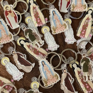 May include: A collection of colourful keychains featuring religious figures and images. The keychains are made of clear acrylic and have a silver metal ring. The figures include a woman in a blue dress, a man in a red robe, and a woman in a white dress.