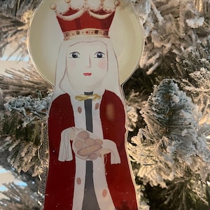 May include: A clear, circular Christmas ornament depicting a stylized figure with a red crown and robe, holding a bundle. The ornament hangs on a snow-covered Christmas tree. The text "Elizabeth of Hungary" is visible.