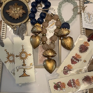 Puede incluir: Una colecci&oacute;n de artesan&iacute;as y decoraciones de tem&aacute;tica religiosa, que incluye un coraz&oacute;n dorado con una cruz, una imagen enmarcada de una estrella dorada y varios marcadores con im&aacute;genes religiosas. Los art&iacute;culos est&aacute;n dispuestos sobre una superficie blanca con un marco decorativo.