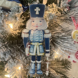 Puede incluir: Un adorno de cascanueces azul y blanco con ribetes dorados y una cruz en su sombrero. Está sosteniendo una espada en su mano derecha.