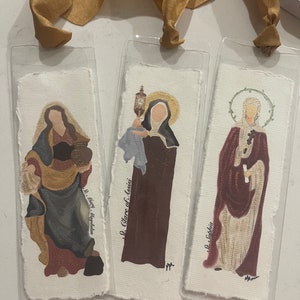 Może przedstawiać: Trzy zakładki z obrazami świętych. Zakładki są wykonane z papieru i zamknięte w przezroczystym plastiku. Pierwsza zakładka przedstawia obraz Świętej Marii Magdaleny, druga zakładka przedstawia obraz Świętej Klary z Asyżu, a trzecia zakładka przedstawia obraz Świętej Zofii.
