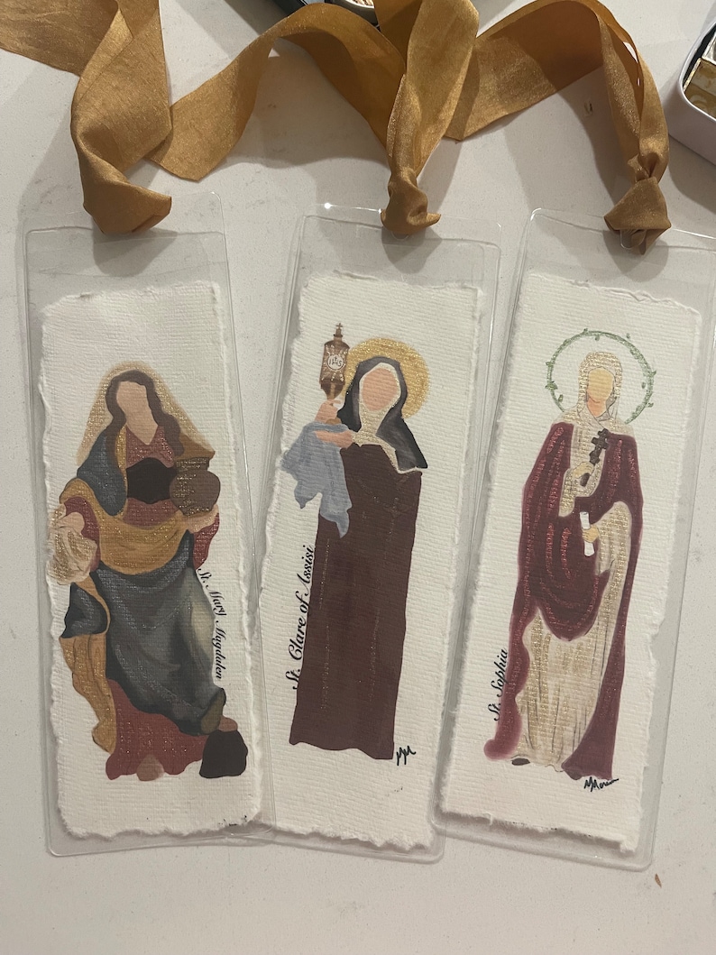 Może przedstawiać: Trzy zakładki z obrazami świętych. Zakładki są wykonane z papieru i zamknięte w przezroczystym plastiku. Pierwsza zakładka przedstawia obraz Świętej Marii Magdaleny, druga zakładka przedstawia obraz Świętej Klary z Asyżu, a trzecia zakładka przedstawia obraz Świętej Zofii.