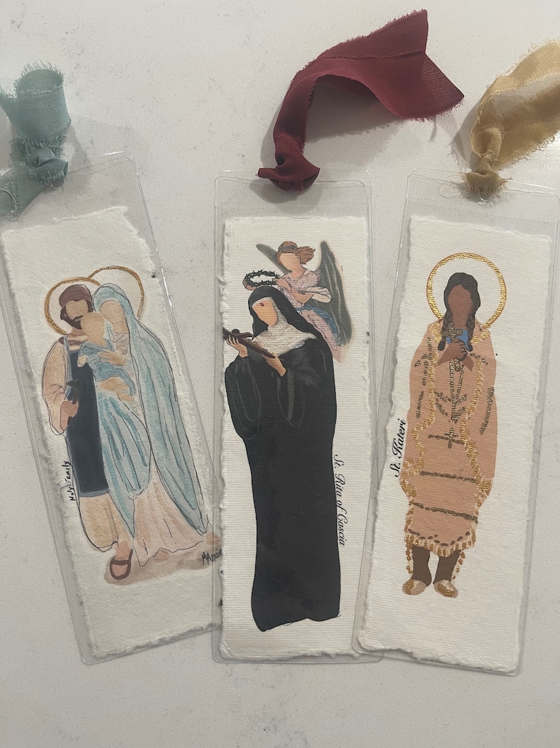 Może przedstawiać: Trzy ilustrowane zakładki z postaciami religijnymi. Pierwsza zakładka przedstawia Świętą Rodzinę, druga zakładka przedstawia Świętą Ritę z Cascii, a trzecia zakładka przedstawia Świętą Kateri.