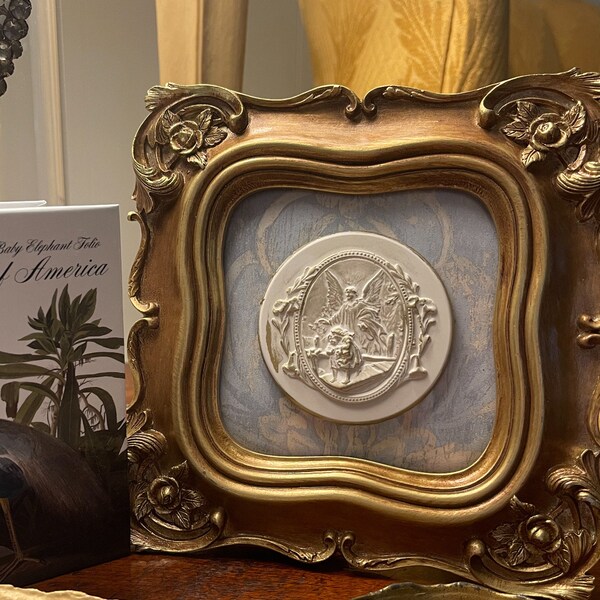 Puede incluir: Una imagen enmarcada en oro con un fondo blanco y un medallón ovalado blanco con una escultura en relieve de un ángel. El ángel sostiene una cruz y está rodeado de una corona de flores. La imagen está sobre una mesa de madera con otros objetos decorativos.