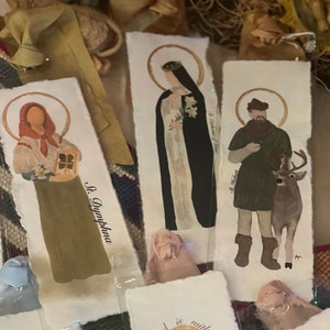Może przedstawiać: Zestaw pięciu ilustrowanych zakładek z postaciami religijnymi i tekstem. Zakładki są wydrukowane na białym papierze o brązowej fakturze. Zakładki mają r&oacute;żne rozmiary i kształty. Zakładki są ułożone jedna na drugiej.