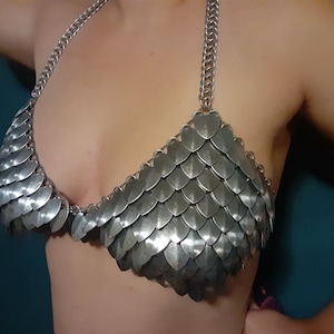 May include: A silver chainmail bra with a scale pattern. The bra is made of small, interlocking metal rings.