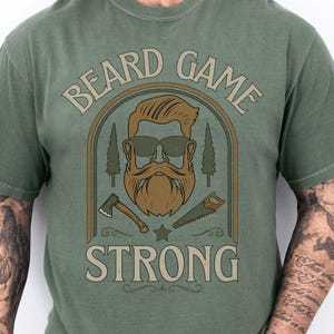 May include: Moss green t-shirt with the words "BEARD GAME STRONG" in a circular design. The design includes a bearded man wearing sunglasses, trees, an axe, and a saw. The shirt is a comfort color.