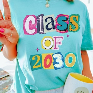 May include: A light blue t-shirt with the text "Class of 2030" in colorful, block letters. A white mug with a yellow interior and a smiley face graphic is held in the hand. The shirt is a light blue color.