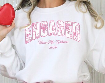 May include: White sweatshirt with the word "ENGAGED" in pink floral lettering. Below the word "ENGAGED" is the text "Future Mrs Williams 2026". A red heart-shaped wallet is held in the left hand. The sweatshirt is part of the "So Pink Custom" collection.