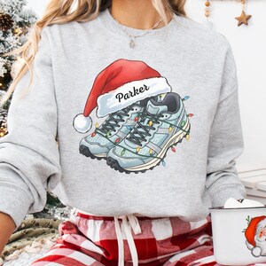 May include: Ash gray sweatshirt with a festive design. The graphic features a pair of light blue running shoes adorned with Christmas lights and a Santa hat with the name "Parker." The model is wearing red and white plaid pajama pants. A mug with a Santa Claus design is in the model's hand.