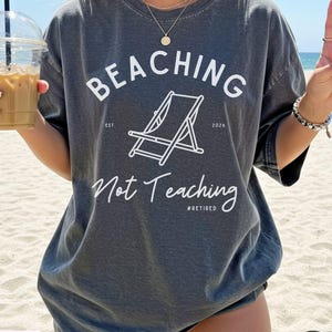Comfort Colors Shirt with deck chair illustration in the center, curved text above, script below - BEACHING Not Teaching #Retired Year Est. 2026