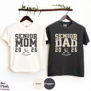 May include: Two t-shirts, one ivory and one pepper, hanging on a wooden rod. The ivory shirt reads "Senior Mom 2026" with a golf ball and club graphic. The pepper shirt reads "Senior Dad 2026" with a similar graphic. Both shirts have the name "Alexander" below.