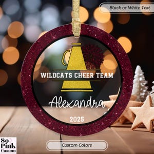 May include: Round ornament with a maroon glitter border and a gold ribbon for hanging. The ornament features a gold megaphone and pom-pom design with the text "WILDCATS CHEER TEAM" and the name "Alexandra" and the year "2025".