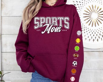 May include: Maroon hoodie with the text "SPORTS Mom" in white, with "EST. 2010" below. The left sleeve features sports ball graphics and names. The hoodie has a front pocket and a hood.