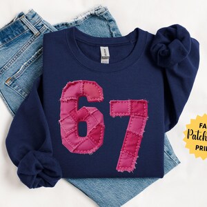 May include: Navy blue sweatshirt with a pink patchwork design of the number 67. The sweatshirt is folded over blue jeans. The words "Faux Patchwork Print" are in a yellow starburst. The words "So Pink Custom" are in the lower left corner.