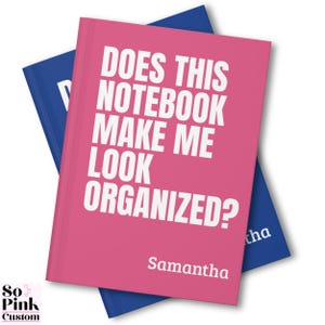 May include: Two notebooks, one pink and one blue, are stacked. The pink notebook has the text "DOES THIS NOTEBOOK MAKE ME LOOK ORGANIZED?" in white, with the name "Samantha" below. The blue notebook is partially visible.