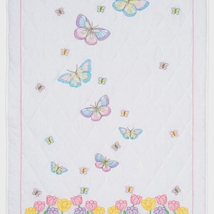 Puede incluir: Manta acolchada blanca con mariposas bordadas en colores pastel. La manta presenta un borde de tulipanes bordados en amarillo, rosa y morado. Un borde rosa enmarca la manta.