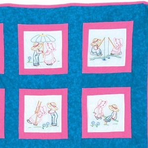 Puede incluir: Un edredón hecho a mano con fondo azul y ribete rosa. El edredón presenta cuadrados bordados con escenas de dos figuras, posiblemente animales, participando en diversas actividades. Los cuadrados están enmarcados en rosa.