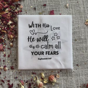 May include: A white square cloth printed with the words "WITH His Love He will calm all YOUR FEARS" in black script, with a heart and stars. The scripture reference Zephaniah 3:17 is at the bottom. Dried flower petals are scattered around the cloth.