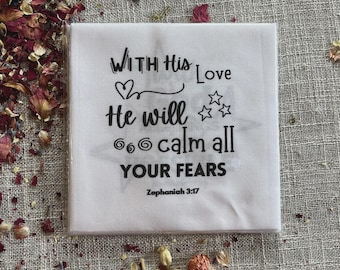 Könnte beinhalten: Ein quadratisches, weißes Tuch mit dem schwarzen Text "WITH His Love He will calm all YOUR FEARS", verziert mit einem Herz und Sternen. Der Bibelvers Zephanja 3:17 steht unten. Getrocknete Blütenblätter umgeben das Tuch.