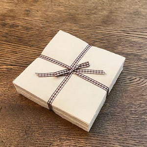 Puede incluir: Una pila de tarjetas cuadradas de papel beige atadas con una cinta a cuadros marrón y blanca. Las tarjetas están cuidadosamente apiladas y la cinta está atada con un lazo. El fondo es una superficie de madera oscura.