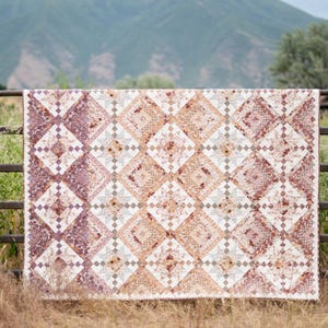 May include: A handmade quilt with a geometric pattern in shades of mauve, peach, and cream hangs on a metal gate. The quilt's design features diamond shapes and floral accents. The background includes a mountain and grassy field.