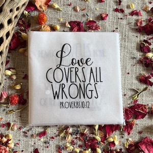 May include: A white square cloth with the text "Love COVERS ALL WRONGS PROVERBS 10:12" in black script. The cloth is surrounded by dried red flower petals and other natural elements.