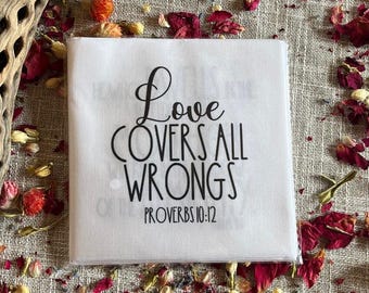 Könnte beinhalten: Ein weißes quadratisches Tuch mit dem Text "Love COVERS ALL WRONGS PROVERBS 10:12" in schwarzer Schrift. Das Tuch ist von getrockneten roten Blütenblättern und anderen natürlichen Elementen umgeben.