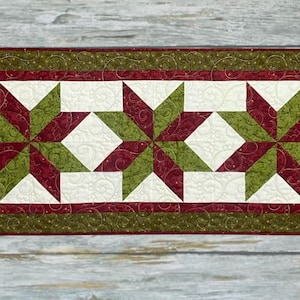 Peut inclure: Un chemin de table matelassé rectangulaire présentant trois motifs d'étoiles à huit branches dans des tons alternés de rouge foncé et de vert olive. Les étoiles sont sur un fond crème avec des motifs de surpiqûres en spirale et une bordure vert foncé.
