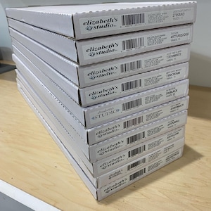 May include: A stack of white cardboard boxes with the "elizabeth's studio" logo. Each box has a label with text including color names like "Sand", "Turquoise", "White", "Cream", "Black", "Purple", and "Yellow".