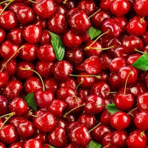May include: A close-up of a pile of ripe cherries. The cherries are a deep red colour, with some showing water droplets. Green leaves are scattered throughout, and the stems are visible.