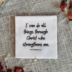 May include: A white square textile with the text "I can do all things through Christ who strengthens me. Philippians 4:13" in black. The textile is on a textured, neutral-colored surface, with dried flowers scattered around.