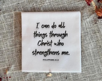 Könnte beinhalten: Ein weißes quadratisches Textil mit dem Text "I can do all things through Christ who strengthens me. Philippians 4:13" in Schwarz. Das Textil liegt auf einer strukturierten, neutralfarbenen Oberfläche, mit getrockneten Blumen verstreut.