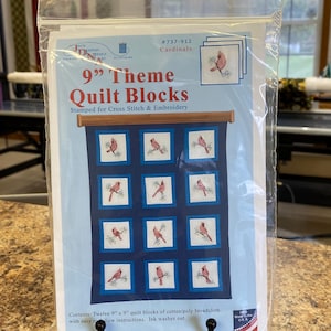 May include: A clear plastic package containing 9-inch theme quilt blocks for cross stitch and embroidery. The label features the text 9" Theme Quilt Blocks, Cardinals, and #737-912, showing twelve white fabric squares with red cardinal bird designs.