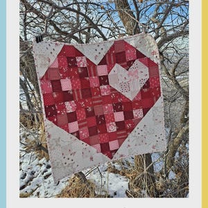May include: A heart-shaped quilt made of red, pink, and white fabric squares. The quilt is hanging from a tree branch. The words "Scrappy Love" are at the top. The Aunt Em's Quilts logo is at the bottom.
