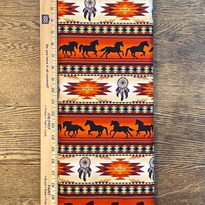 May include: Orange, brown and blue fabric with a repeating pattern of Native American inspired geometric designs, dreamcatchers, and silhouettes of horses. The fabric is measured with a ruler in inches.