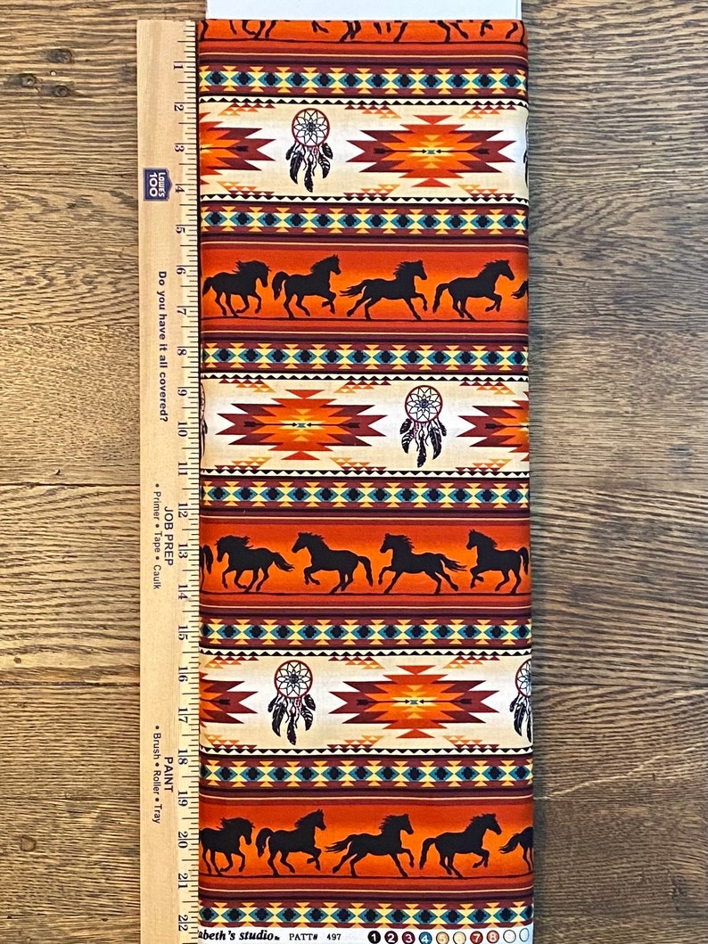May include: Orange, brown and blue fabric with a repeating pattern of Native American inspired geometric designs, dreamcatchers, and silhouettes of horses. The fabric is measured with a ruler in inches.